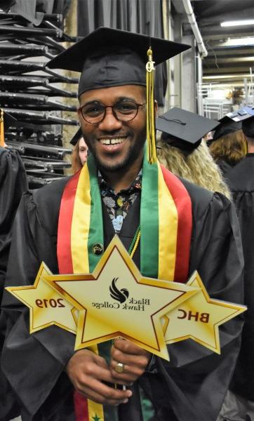 Amadou Ndiaye wearing graduation gown. Holding 3 signs shaped like gold stars saying "BHC", "bbin信誉网站", and "2019"