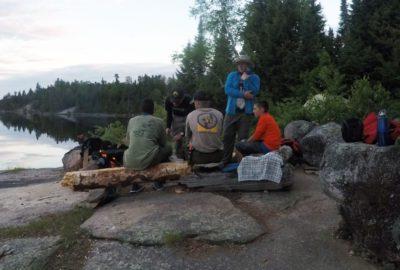 a group of scouts around a campfire on the shore of a lake