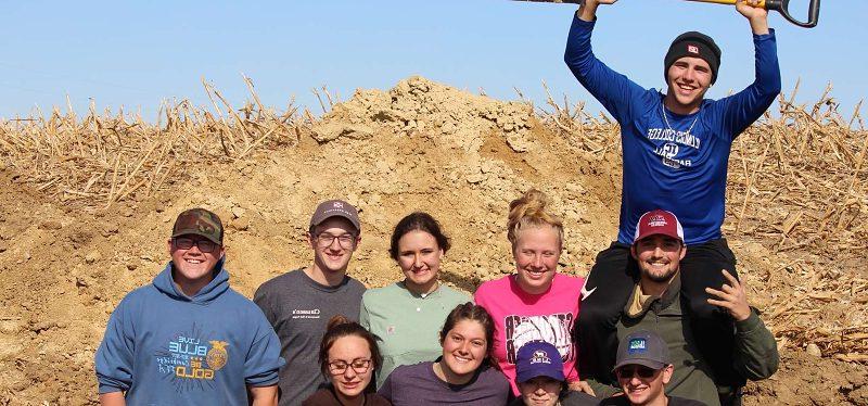 10 students in soil pit with one holding shovel above head