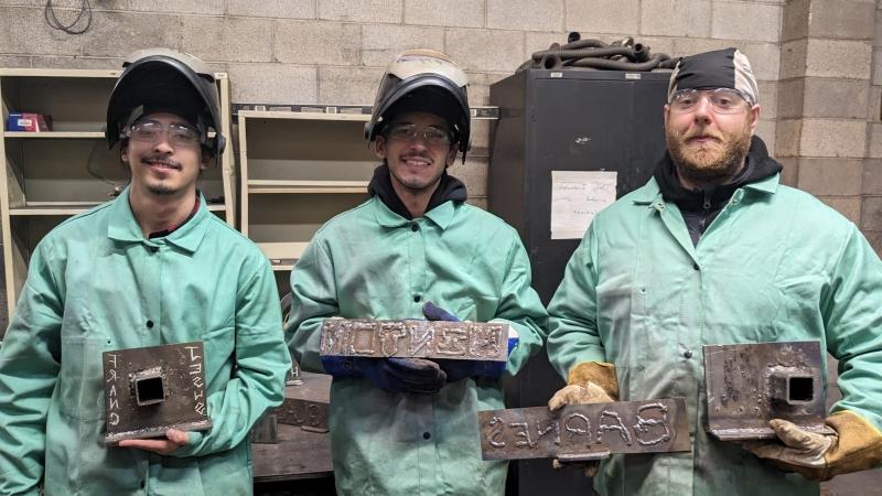 3 students in welding gear holding up nameplates they welded